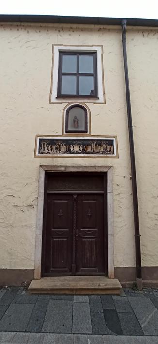 Quadratisches Fenster mit mittigem Festerkreuz über Tür und Heiligenkasten. Dieser Heilige ist auch hinter Glas, dieses Fenster hat die Form einer Tür mit Rundbogen. Darunter sieht man einen Kasten mit Inschrift "Antoni Silcher - 1864 Antoni-Bräu!", bevor dann die dunkel Tür kommt. Das Haus ist in hellem Lindgrün gestichen, rechts der Türen und Fenster verläuft eine braune Dachrinne von oben nach unten.