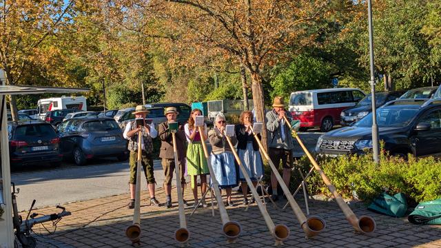 Sechs Alphornbläser spielen vor herbstlich gefärbten Bäumen in oberbayerischer Tracht.