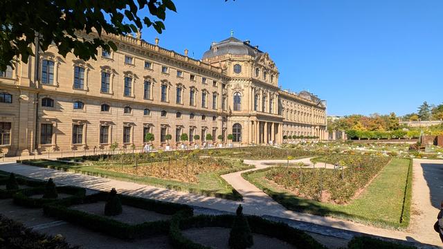 Zweistöckiges Gebäude mit Mittelkuppel im barocken Stil mit großen Garten vor dem Gebäude. Gebäude aus hellem ockerfarbenem Sandstein, hohen fenstern und einer Balustrade au fdem Dach. Garten mit Blumenbeeten und Kieswegen angelegt in beginnenden Herbstfarben. Sonnenlicht um die Mittagszeit bei strahlend blauem Himmel.