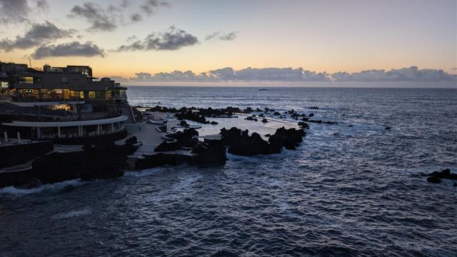 Blick auf Felsenküste im Abendlicht, ruhiger Wellengang und links ragen Felsenpools und ein schwach beleuchtetes Hotel ins Bild. Es wird von hinten mit orangem Himmel beleuchtet, an dem über dem Wasser graublauen Wölkchen stehen.